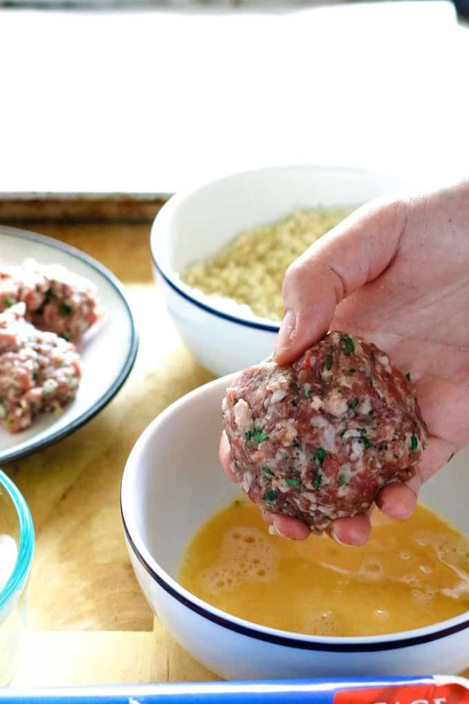 egg wrapped in meat being dipped into beaten egg