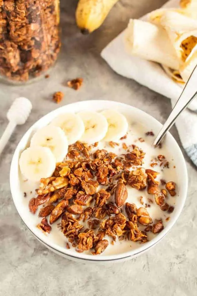 white bowl full of homemade honey nut granola and almond milk with sliced bananas on a concrete countertop