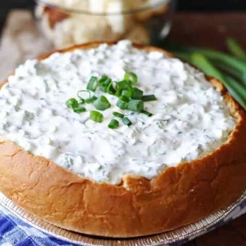 A delicious green onion dip served in a hollowed-out round loaf of freshly baked bread. The dip is creamy and garnished with finely chopped green onions on top. The bread bowl has a golden crust and is sliced into cubes for dipping. The appetizing presentation is perfect for parties and gatherings.