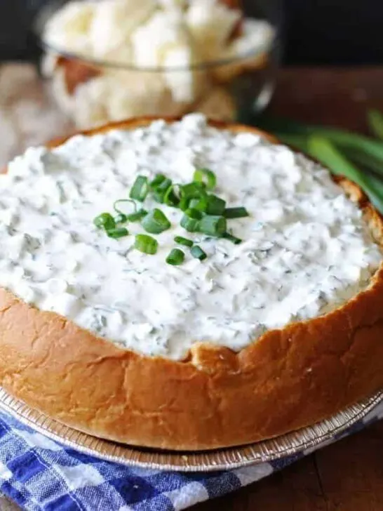 A delicious green onion dip served in a hollowed-out round loaf of freshly baked bread. The dip is creamy and garnished with finely chopped green onions on top. The bread bowl has a golden crust and is sliced into cubes for dipping. The appetizing presentation is perfect for parties and gatherings.