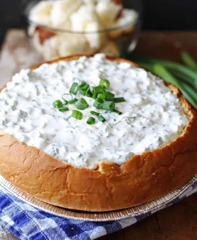 A delicious green onion dip served in a hollowed-out round loaf of freshly baked bread. The dip is creamy and garnished with finely chopped green onions on top. The bread bowl has a golden crust and is sliced into cubes for dipping. The appetizing presentation is perfect for parties and gatherings.