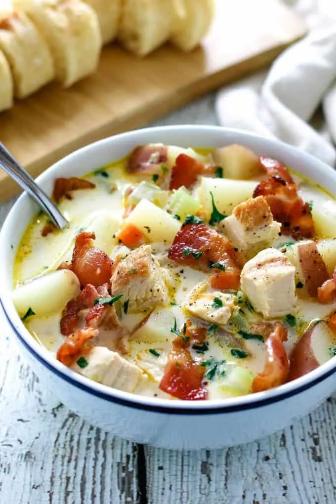 white bowl on a wooden surface. bowl is full of chowder and there is a white napkin and a cutting board of sliced bread