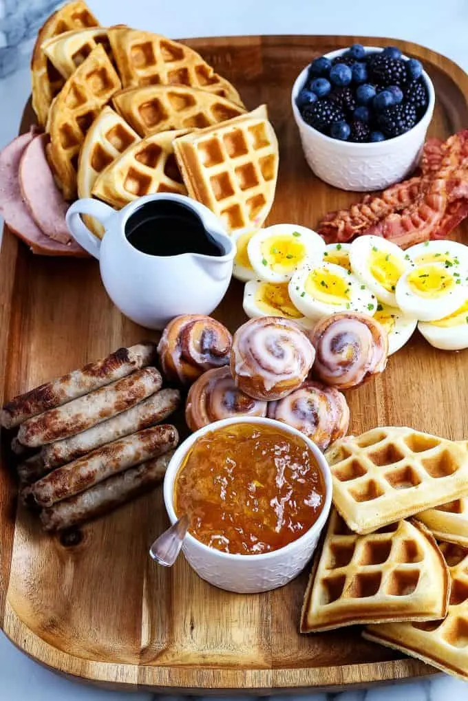 mini cinnamon rolls, deviled eggs, breakfast sausage, waffles, sausage links, Canadian bacon, and berries on a wooden board