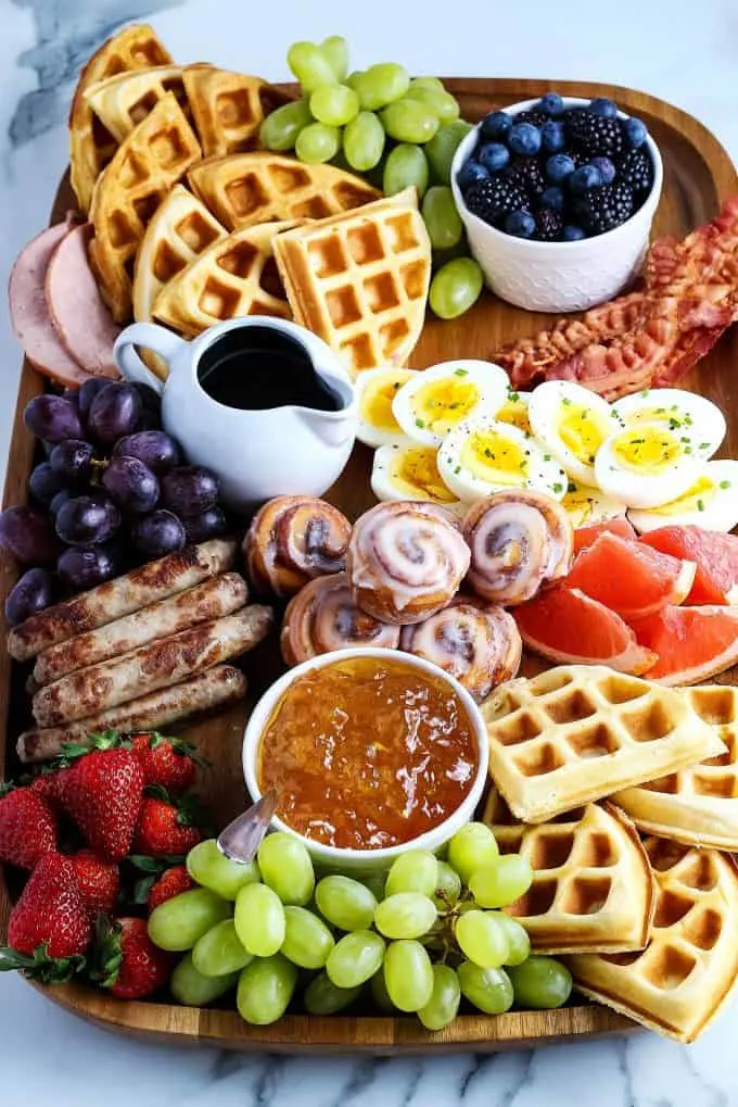 mini cinnamon rolls, deviled eggs, breakfast sausage, waffles, fruit, sausage links, Canadian bacon, and berries on a wooden board