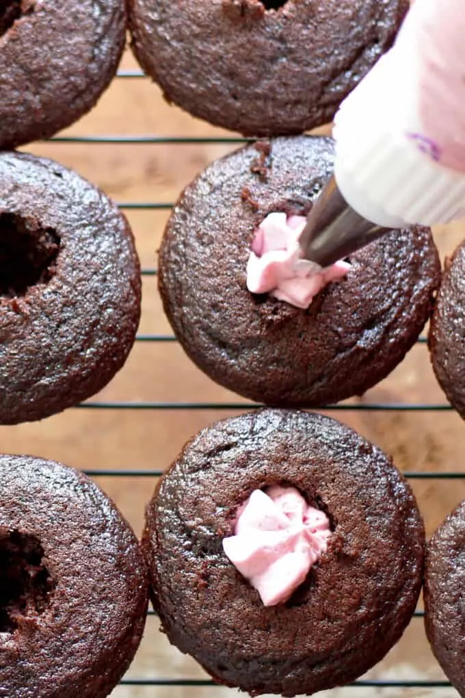 wholes have been punched into cooked chocolate cupcakes. blueberry frosting is in a piping bag and the frosting is being piped into the center of each cupcake