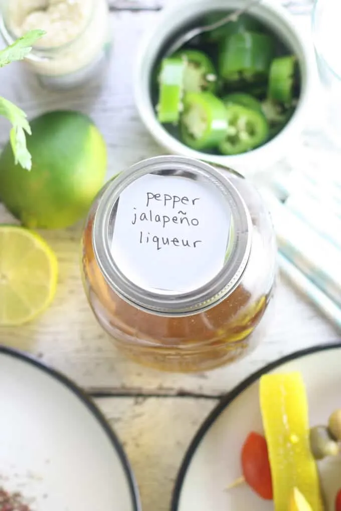 overhead photo of a mason jar with homemade pepper jalapeño liqueur. in the background is a bowl of jalapeño slices and a lime