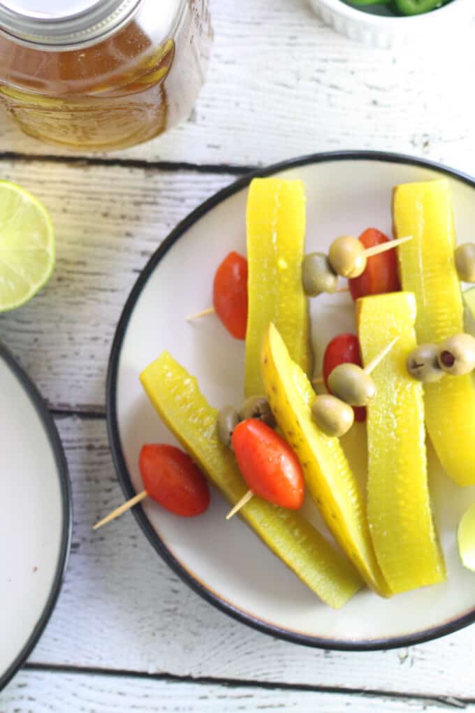 white plate with pre-made Bloody Mary garnishes. the garnishes are on wooden picks and include a cherry tomato, two olives and a pickle spear