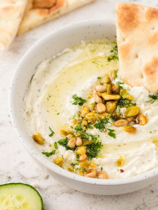 whipped feta dip in a white serving bowl, garnished with pine nuts pistachios and parsley