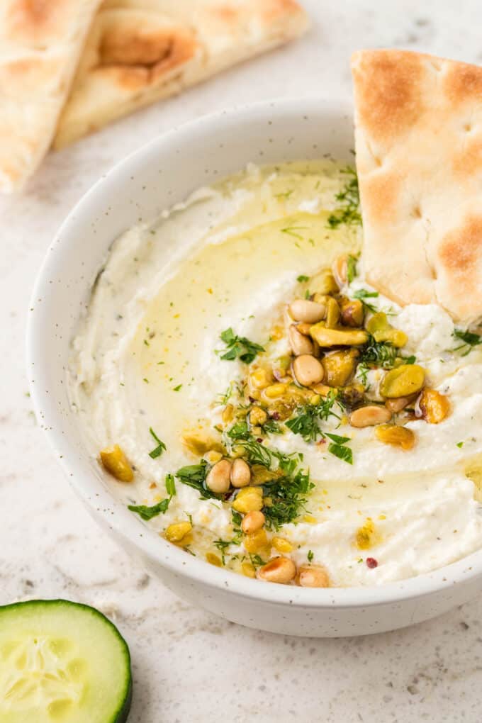 whipped feta dip in a white serving bowl, garnished with pine nuts pistachios and parsley