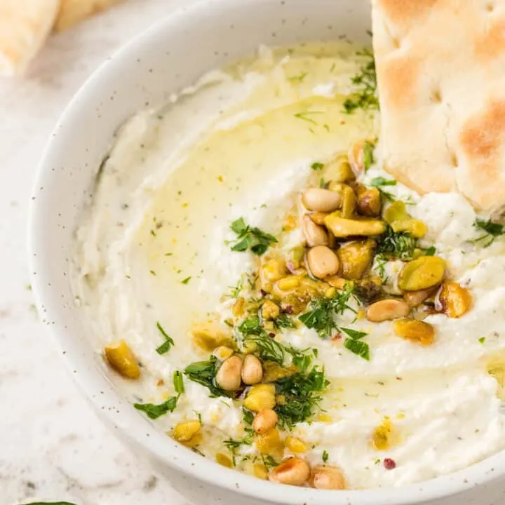 whipped feta dip in a white serving bowl, garnished with pine nuts pistachios and parsley