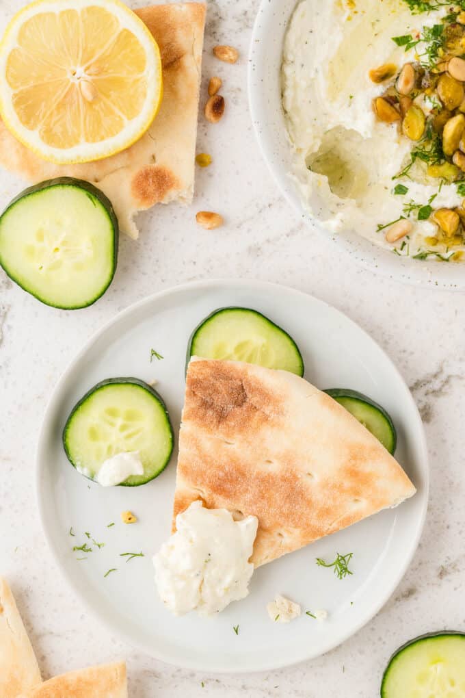 two plates with cucumber slices, lemon wedges, and pita pieces with feta dip on them