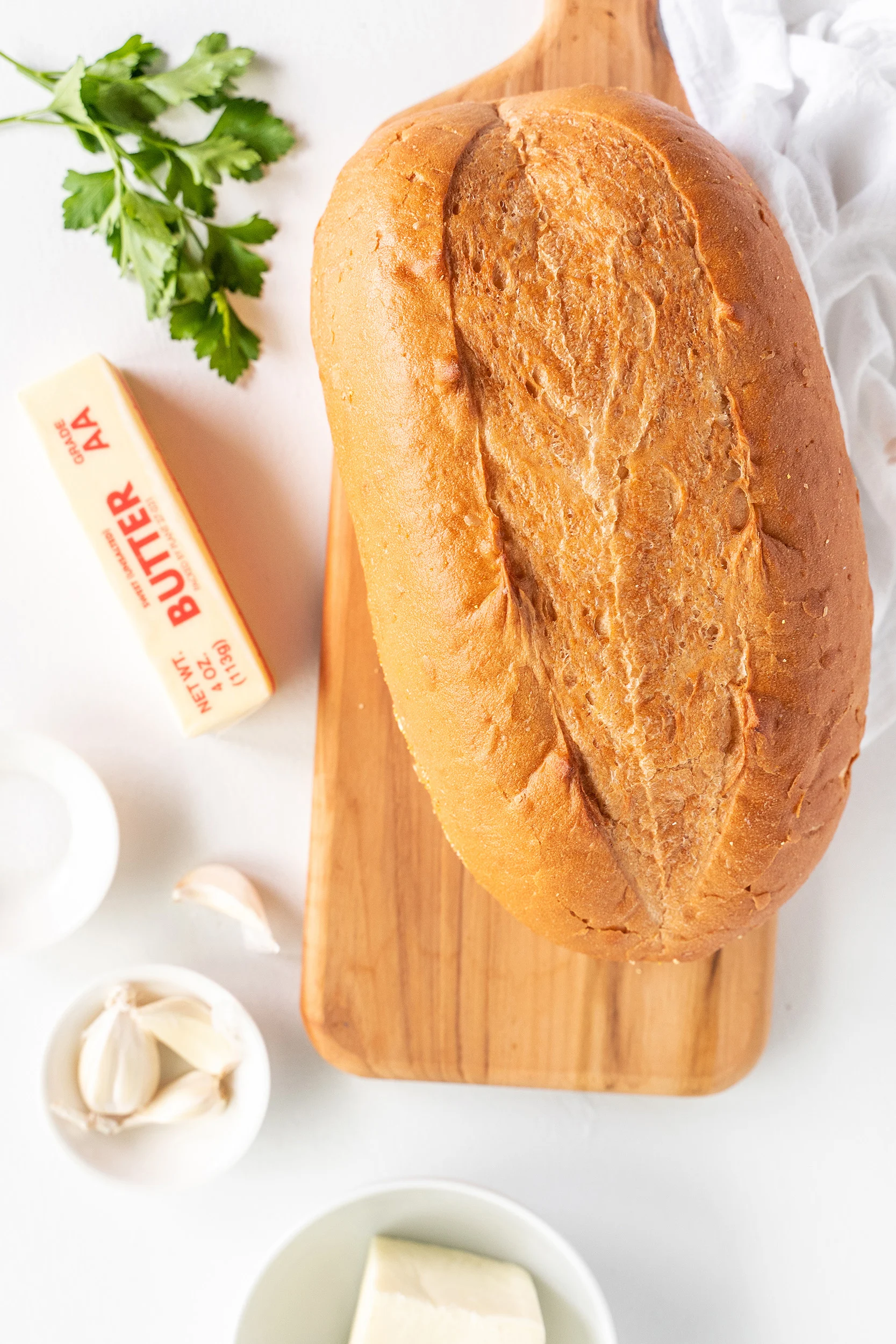 loaf of bread, stick of butter, garlic cloves, cheese, and parsley on a white board