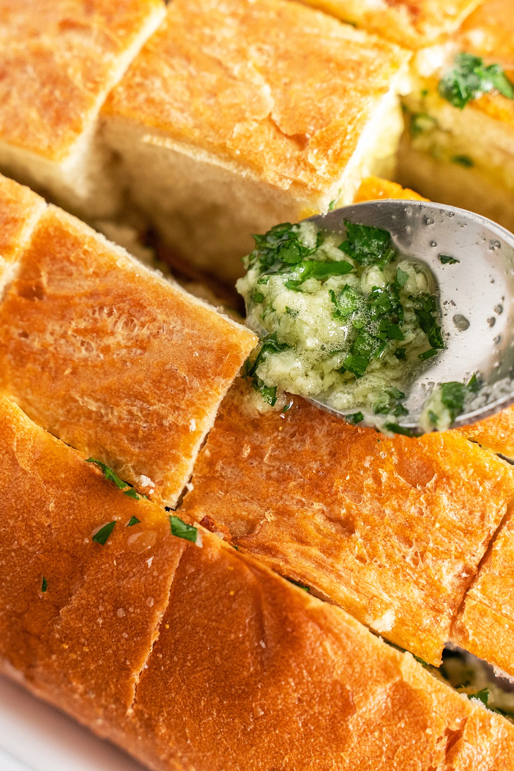 garlic, parsley, and melted butter mixture being spooned in between the cut squares of a loaf of fresh bread