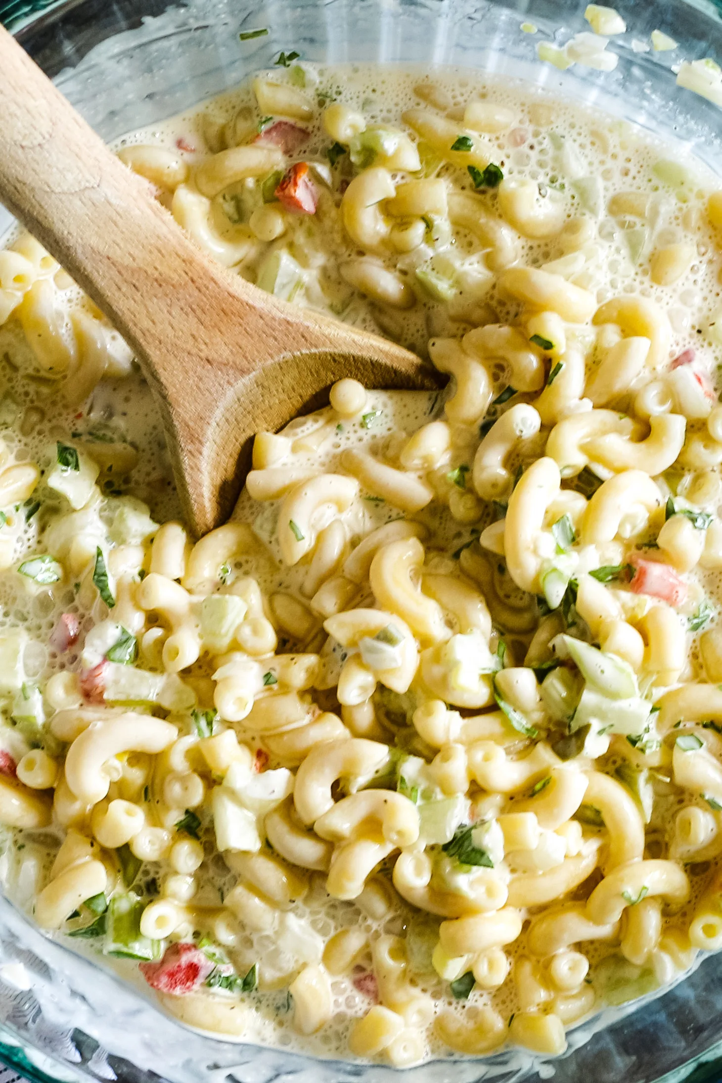 clear mixing bowl with a wooden mixing soon. Inside the bowl is creamy deli macaroni salad