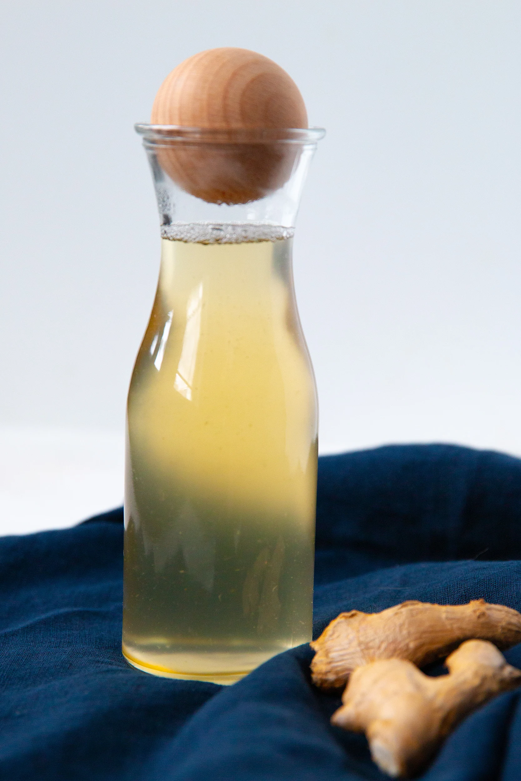 ginger syrup in a tall glass bottle with a round wood stopper. the bottles is on a dark blue towel, with fresh ginger next to it