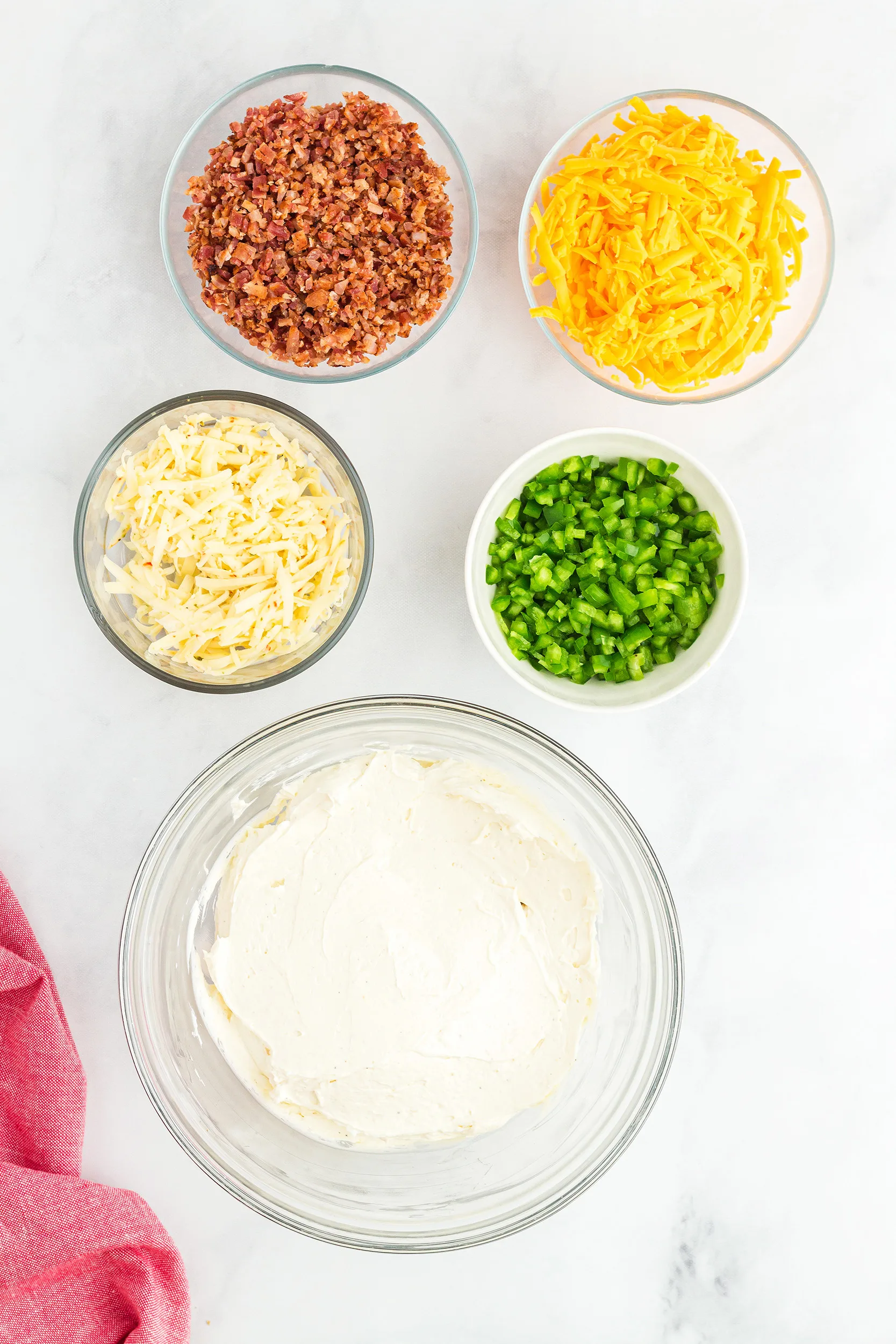 cream cheese and sour cream mixed in a bowl, with shredded cheddar and Havarti cheeses, crispy bacon, and diced jalapeño peppers in small bowls