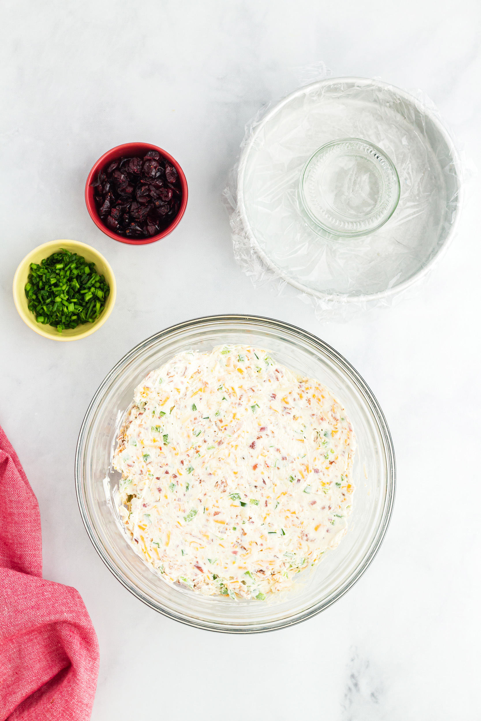 prepared Bacon Jalapeño Cheese Ball ingredients mixed in a bowl with diced chives and dried cranberries next to a plastic wrapped bowl with a glass in the center