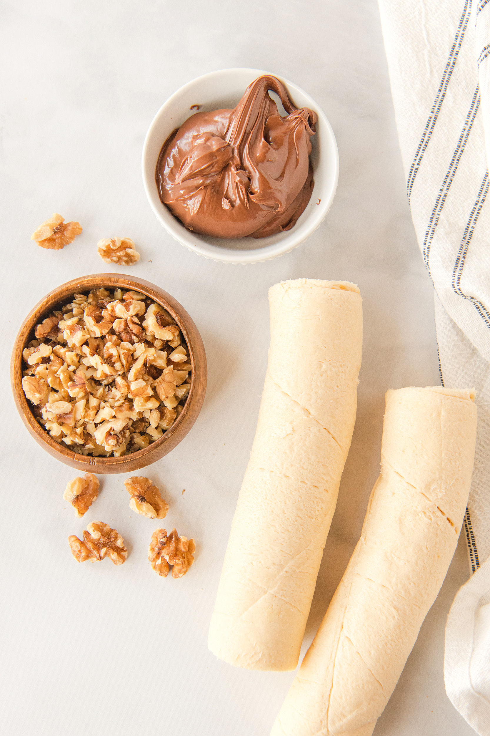 Nutella in a white bowl, walnuts in a wooden bowl, and two packages of crescent roll dough, unpackaged