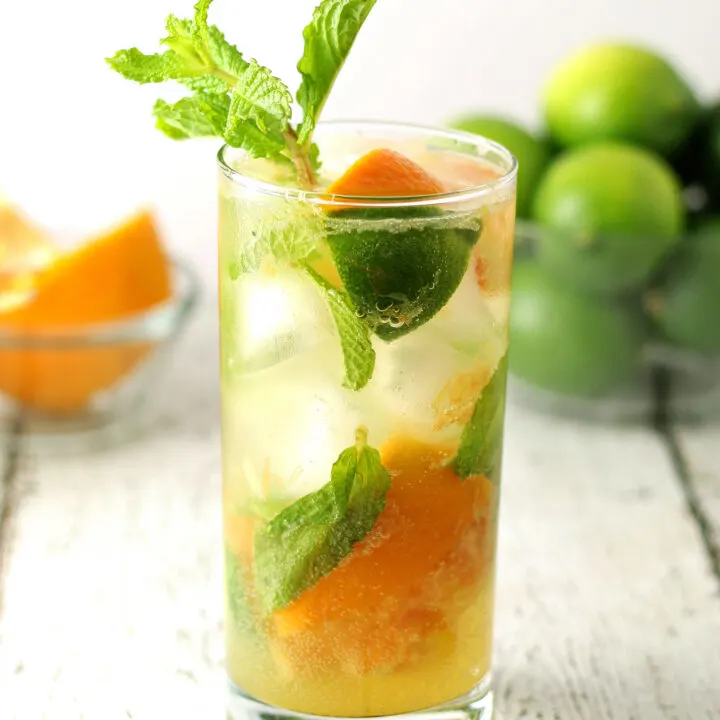 one orange mojito in a clear glass with a bowl of limes and oranges in the background