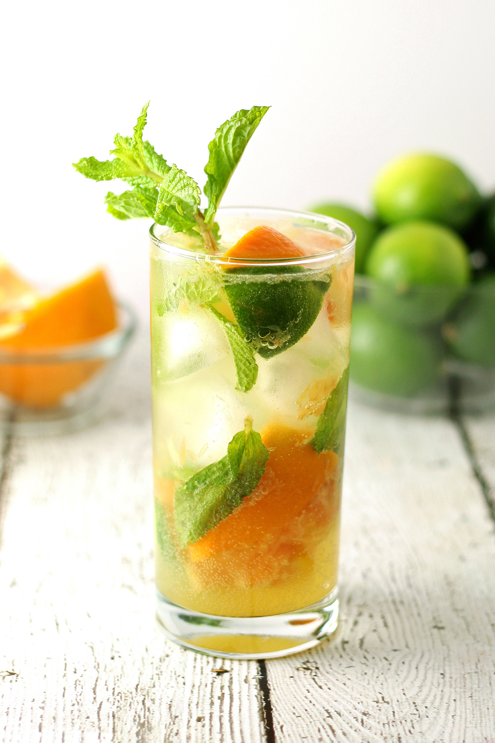 one orange mojito in a clear glass with a bowl of limes and oranges in the background