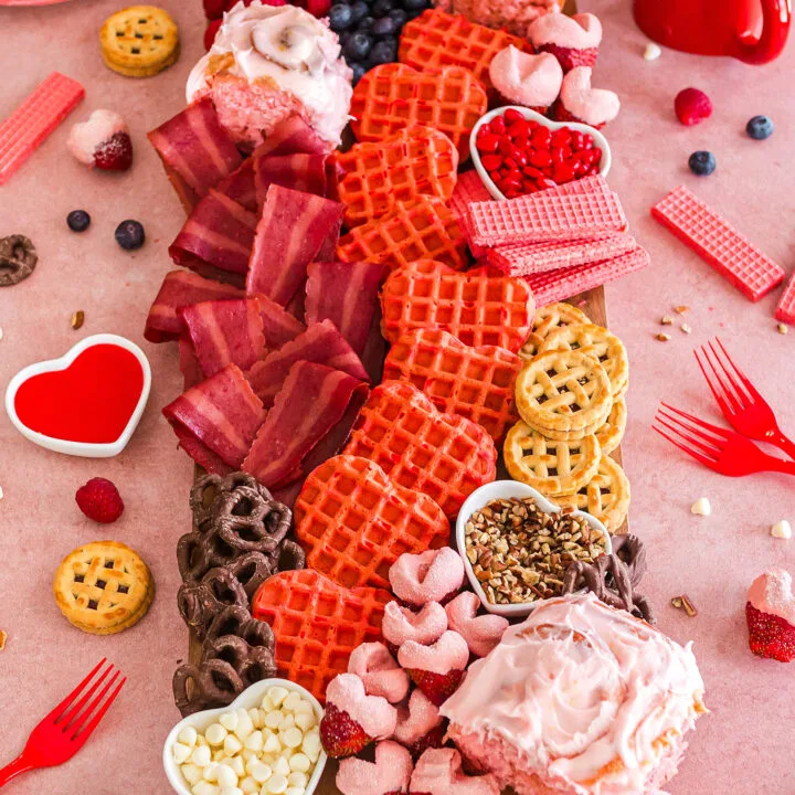 a large wooden board filled with different pink treats including pink heart shaped waffles, strawberry cinnamon rolls, and heart shaped bacon