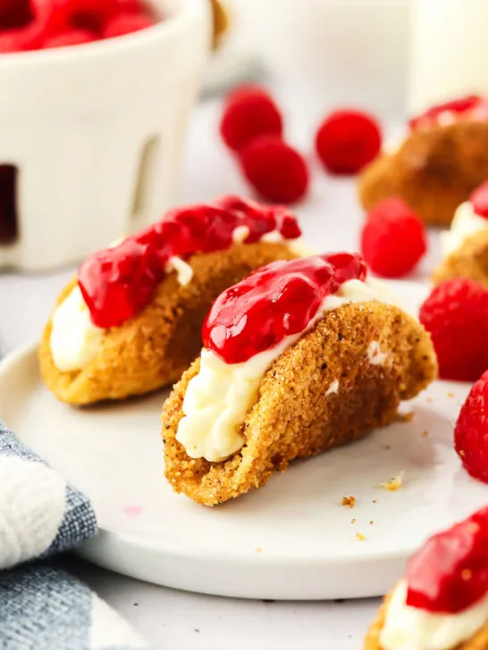 cheesecake tacos on a plate surrounded by fresh raspberries and a blue and white napkin